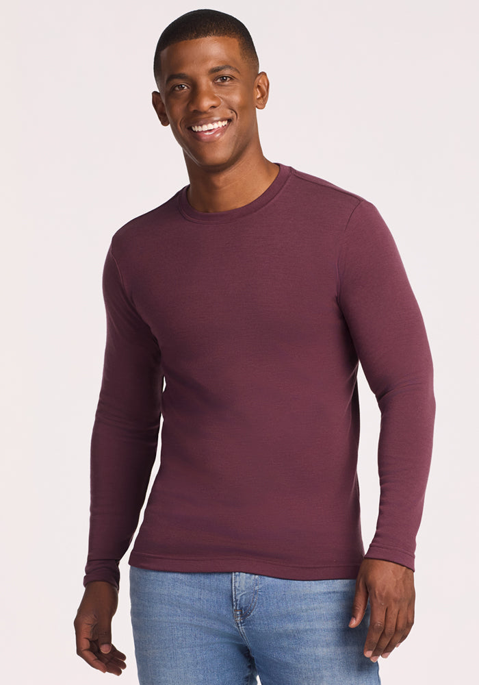 A man smiles while wearing the Woolx Easton Crew Neck Shirt in Grape Harvest and light blue jeans, standing against a plain, light-colored background.