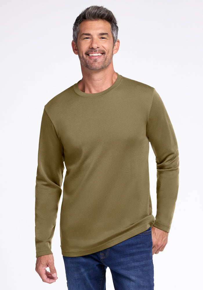 A smiling man with short gray hair and stubble wears the Woolx Easton Crew Neck Shirt in Winter Moss and blue jeans, standing against a plain white background.