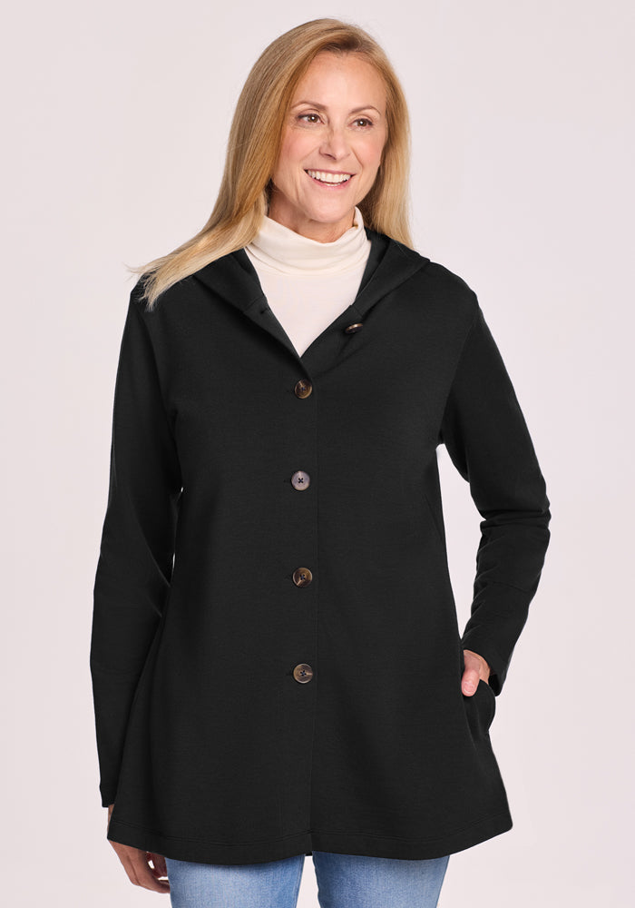 A smiling woman with long blonde hair wears the Woolx Edna Hooded Cardigan in black over a white turtleneck and blue jeans, standing with one hand in her pocket against a light background. | Andrea is 5’10”, wearing a size S