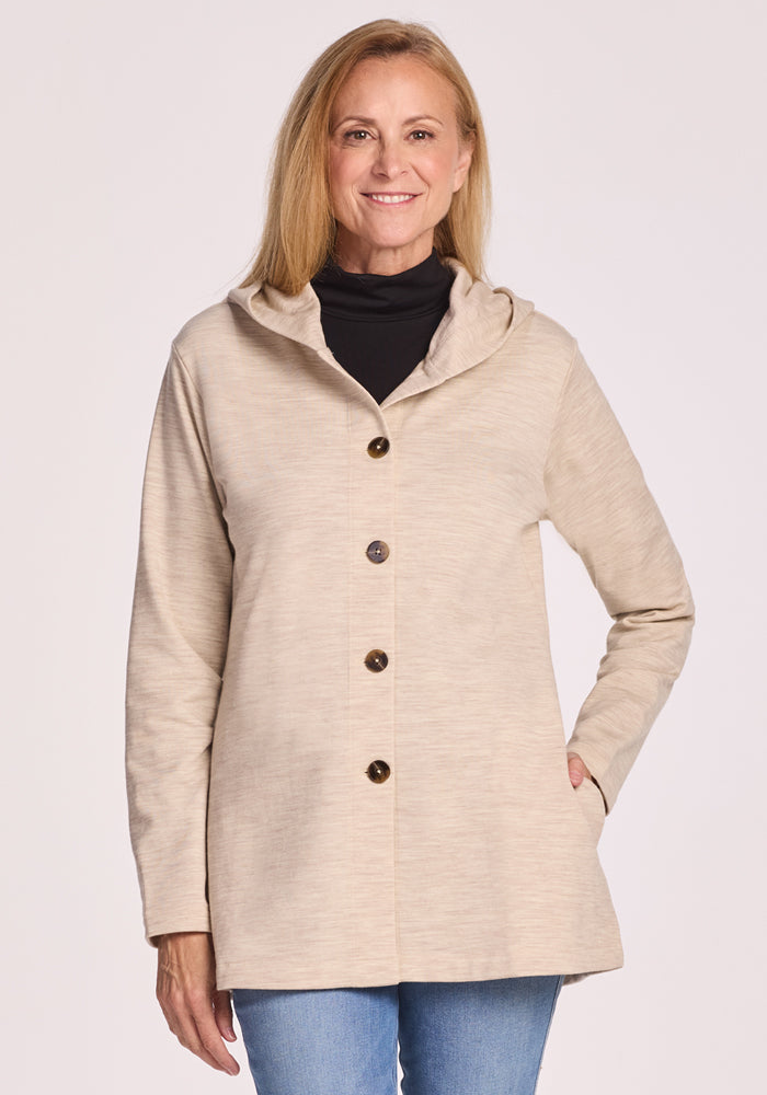 A smiling woman with long blonde hair wears the Woolx Edna Hooded Cardigan in Cream Heather over a black turtleneck and blue jeans, standing against a light background. This cozy piece is perfect for cold weather. | Andrea is 5'10", wearing a size S