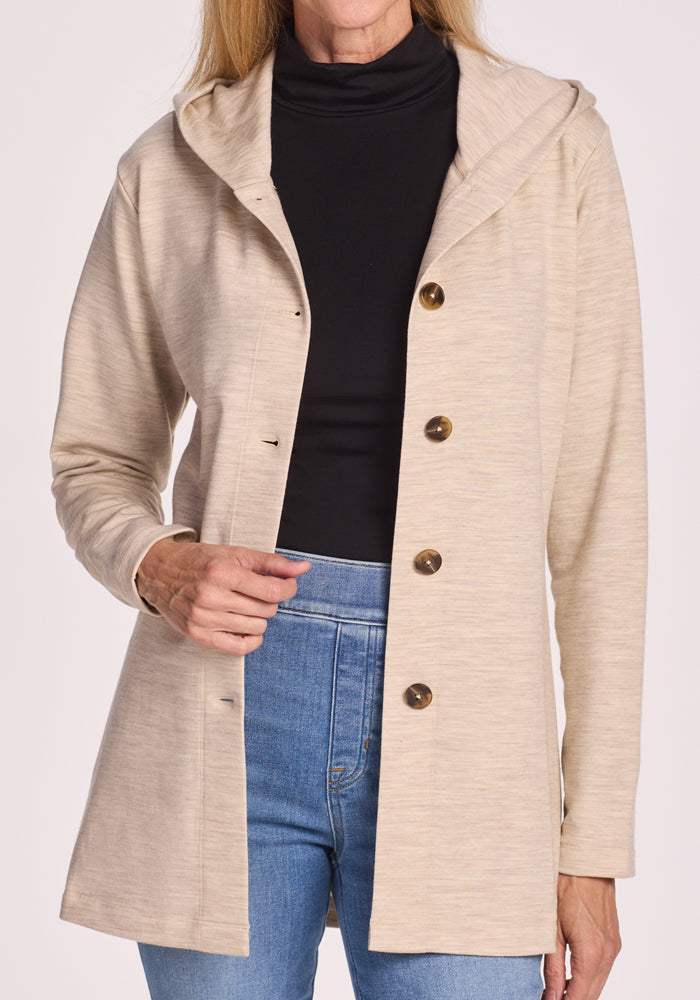 A woman wearing the Woolx Edna Hooded Cardigan in Cream Heather over a black turtleneck and blue jeans stands against a plain light background.