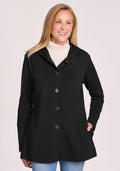 A smiling woman with long blonde hair wears the Woolx Edna Hooded Cardigan in black over a white turtleneck and blue jeans, standing with one hand in her pocket against a light background. | Andrea is 5’10”, wearing a size S