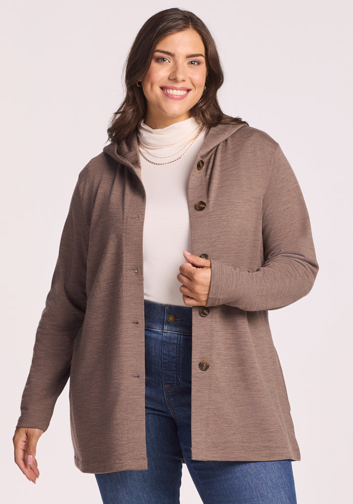 A woman with long brown hair smiles while wearing the Woolx Edna Hooded Cardigan in Simply Taupe, paired with a white turtleneck and blue jeans. She stands against a plain light background.