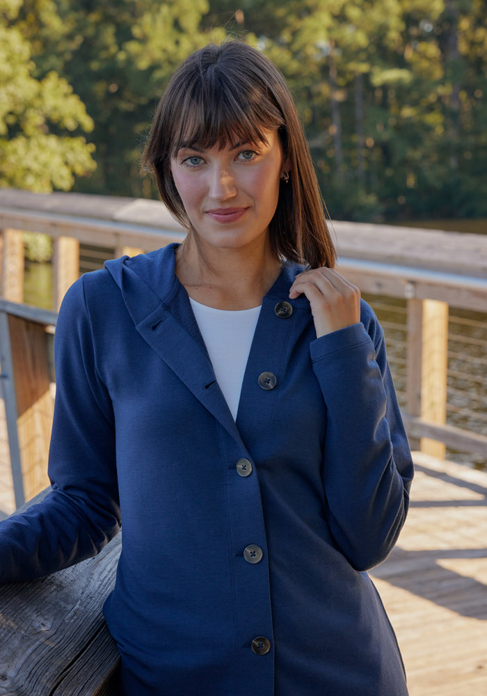 A woman with straight brown hair and bangs stands on a wooden bridge by the water, wearing the Woolx Edna Hooded Cardigan • Final Sale - Deep Navy over a white top. Trees and sunlight can be seen in the background.