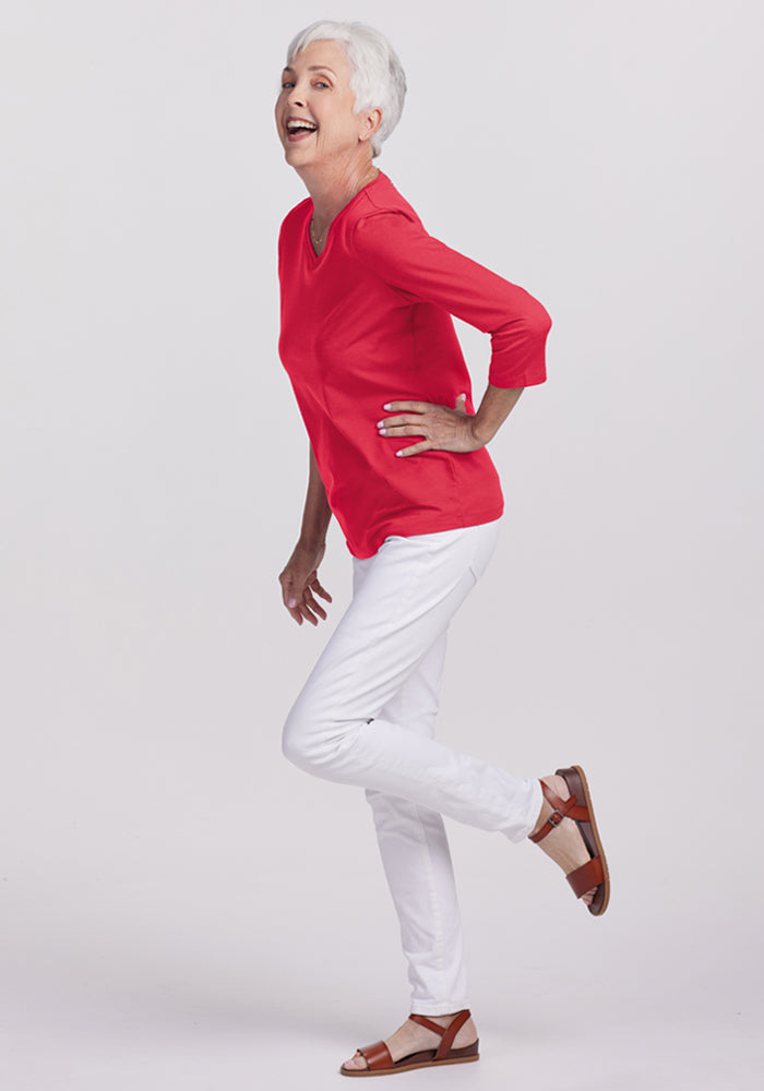 Smiling older woman in a Woolx Elena 3/4 Sleeve V Neck - Berry Ice Pop and white pants, playfully posing on one leg.