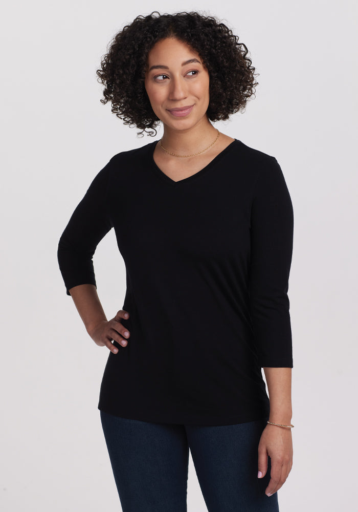 A person with curly hair poses confidently with a hand on their hip, showcasing casual chic in a black Woolx Elena 3/4 Sleeve V-Neck top and dark jeans against a plain white background. | Tori is 5'7", wearing a size S