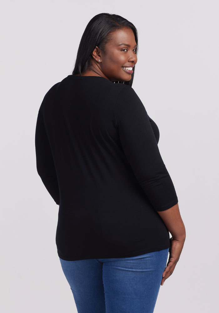 A smiling person with long hair glances over their shoulder, showcasing effortless style in a Woolx Elena 3/4 Sleeve V Neck - Black and blue jeans against a plain, light-colored backdrop.