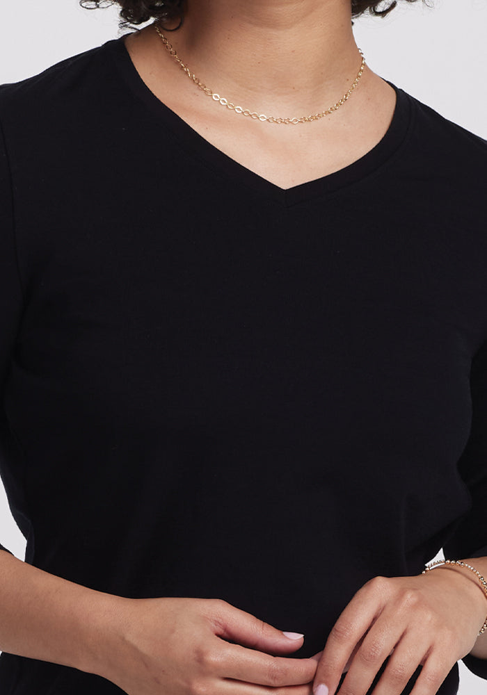 Against a plain background, a person dons the Woolx Elena 3/4 Sleeve V Neck - Black with a delicate gold necklace, arms bent at the elbows and hands gently resting in front, highlighting this versatile merino wool wardrobe essential.
