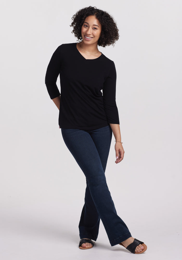A curly-haired person smiles, standing with one foot crossed. They wear a Woolx Elena 3/4 Sleeve V Neck in Black, dark blue jeans, and black sandals, creating a versatile look against the plain white background.