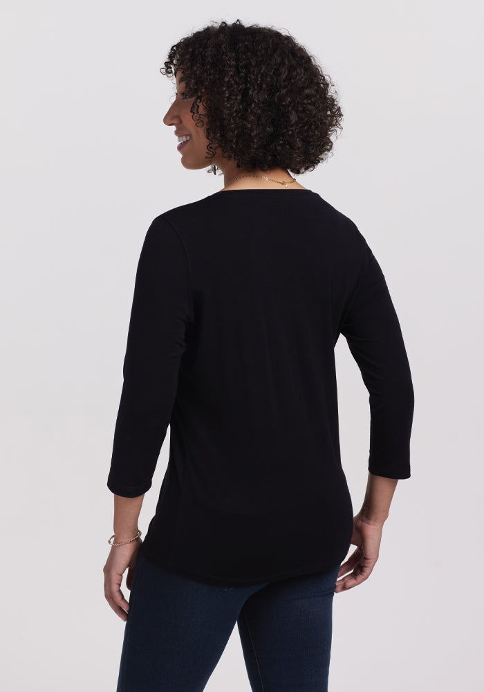 A person with curly hair stands smiling with their head turned to the side against a white background, wearing dark jeans and an Elena 3/4 Sleeve V Neck - Black from Woolx. This merino wool essential merges comfort and timeless style.