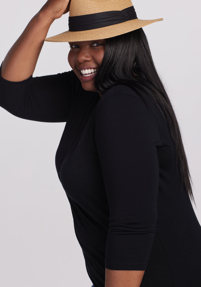 A person in a straw hat with a black band smiles broadly. Their long, straight black hair complements the versatile Elena 3/4 Sleeve V Neck Top in black from Woolx, made of essential merino wool. The background is plain white.