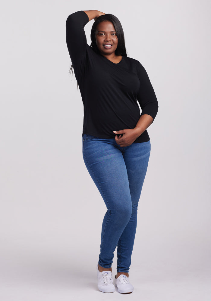 A person with long hair smiles against a plain background, wearing the Woolx Elena 3/4 Sleeve V Neck in black and blue jeans, versatile wardrobe essentials. White sneakers complete the look as one hand rests on their hip and the other touches their head.