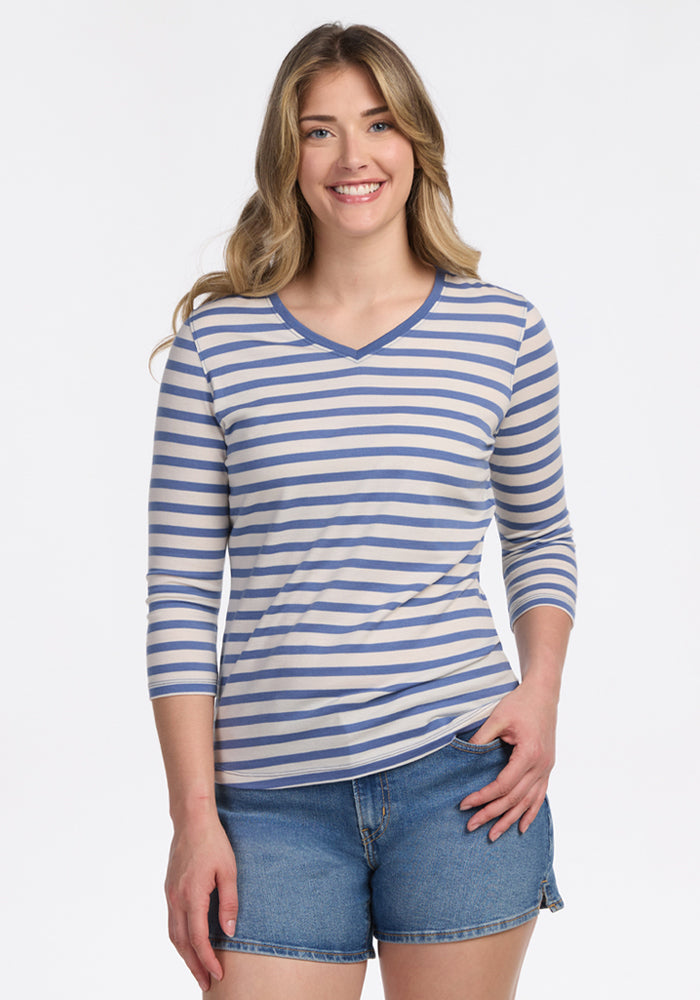 A woman with long, wavy hair smiles at the camera, wearing the Woolx Elena 3/4 Sleeve V Neck in Blue Sand Stripe, paired with denim shorts against a plain white background. | Ariana is 5'11", wearing a size S