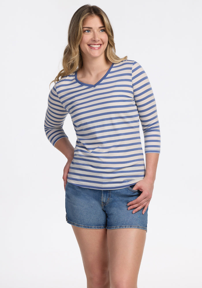 A woman with long, wavy hair smiles in the Woolx Elena 3/4 Sleeve V Neck - Blue Sand Stripe, paired with denim shorts. She stands against a white background, hand on hip, highlighting this stylish and lightweight women’s top.