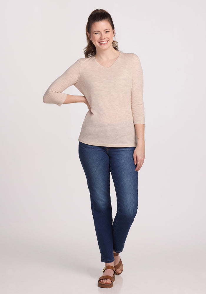 A person with long hair smiles against a plain backdrop, wearing a modern chic Elena 3/4 Sleeve V Neck in Cream Heather by Woolx, dark blue jeans, and sandals. Their right hand is on their hip, and their left arm rests by their side.