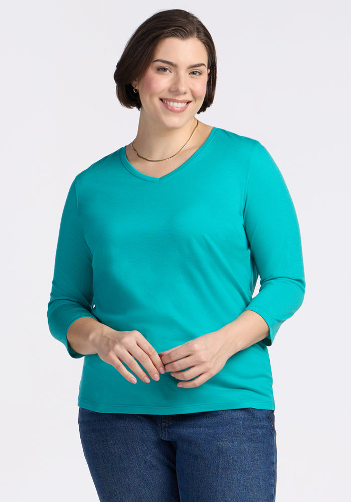 Smiling woman with short brown hair wears Woolx Elena 3/4 Sleeve V Neck in Cool Jade and jeans, plain light background.