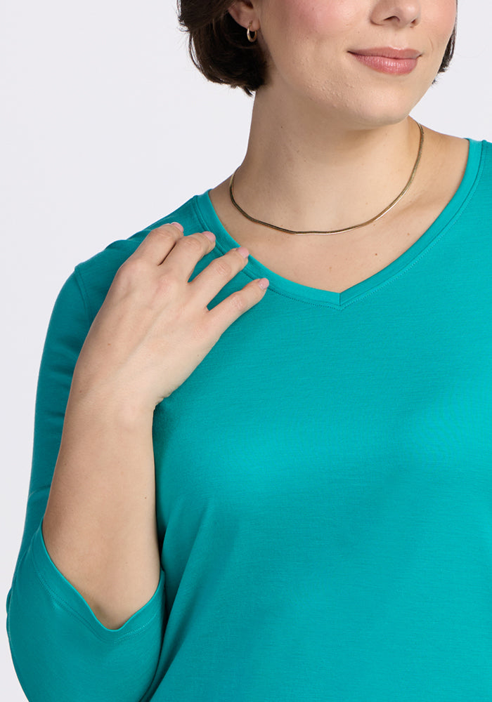Woman in a Woolx Elena 3/4 Sleeve V Neck Top in Cool Jade, wearing a gold necklace and smiling, touches her shoulder.