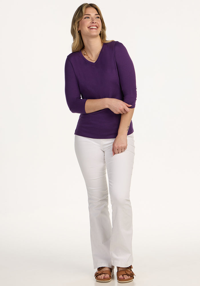 Woman smiling in a Woolx Elena 3/4 Sleeve V Neck - Plum Crush, white pants, and brown sandals against a plain background.