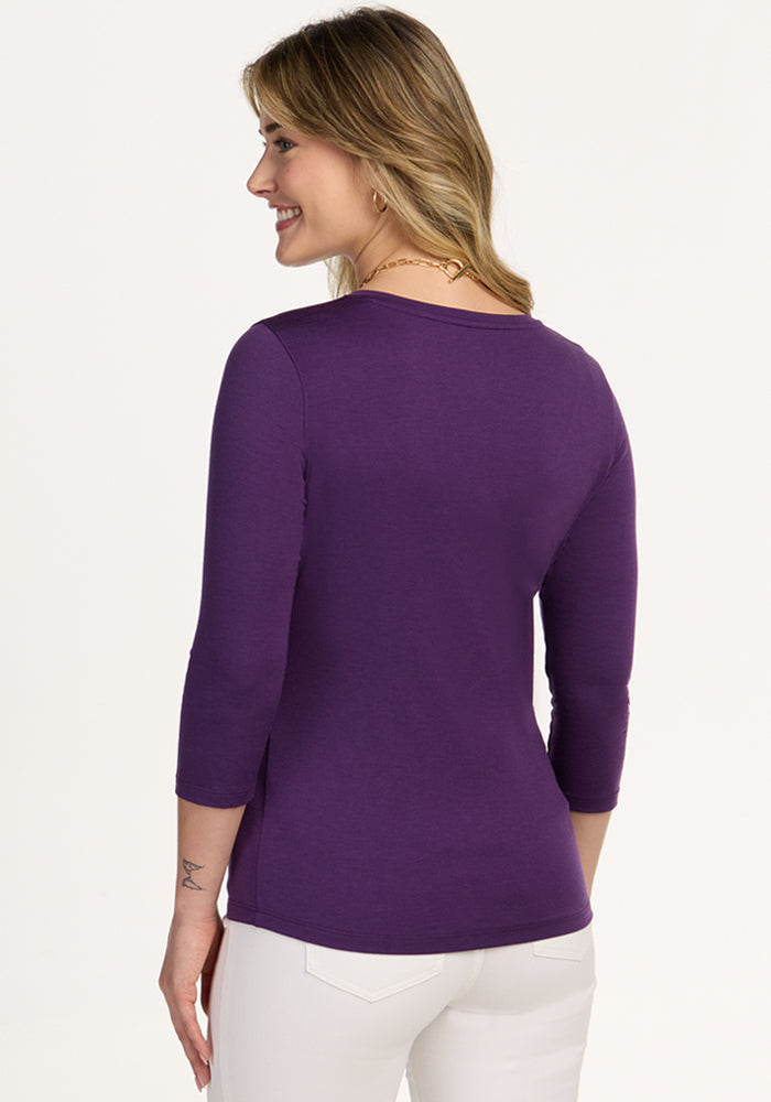 Woman smiles while facing away, wearing Woolx Elena 3/4 Sleeve V Neck in Plum Crush and white pants against a light backdrop.