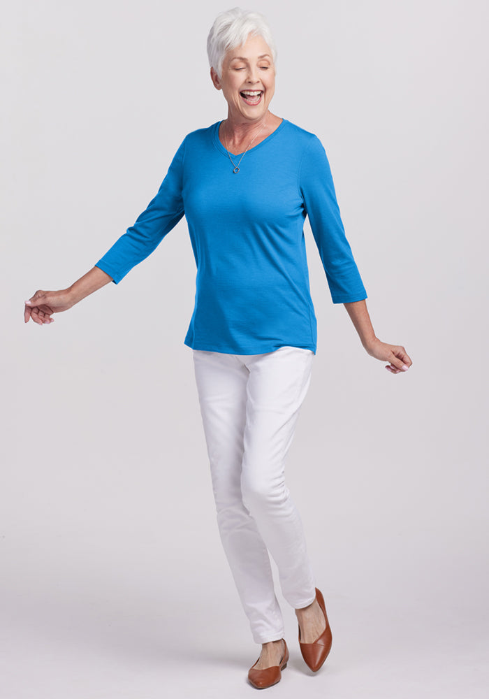 Smiling older woman in a Woolx Elena 3/4 Sleeve V Neck - Wave Rider, white pants, and brown flats poses playfully.