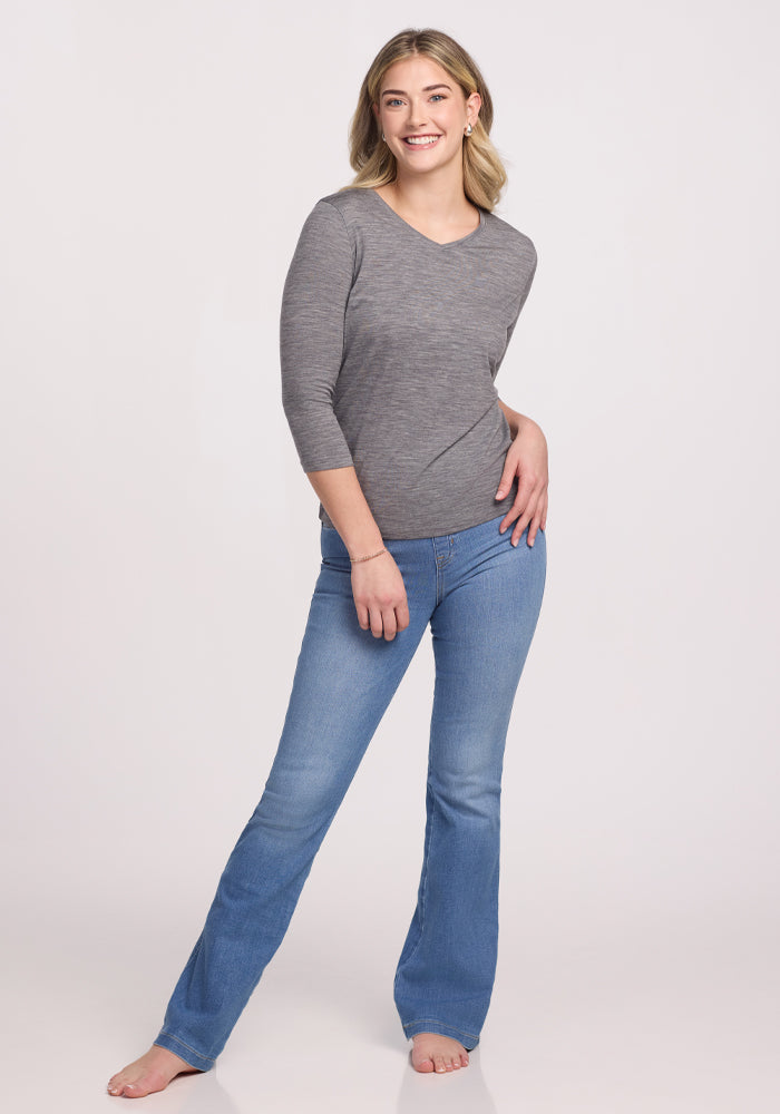 Against a plain backdrop, a barefoot person smiles in relaxed attire, wearing Woolx's sleek Elena 3/4 Sleeve V Neck in Graphite Heather paired with blue jeans. One hand is on their hip while the other casually touches their leg.