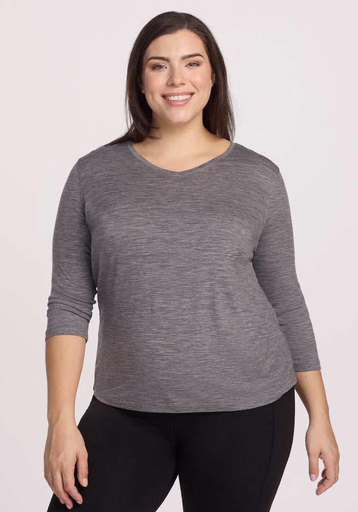 A woman smiles, wearing a Woolx Elena 3/4 Sleeve V Neck in Graphite Heather with black pants, against a light background. The relaxed fit complements her easy stance, arms resting naturally at her sides. | Juliana is 5'10", wearing a size XL