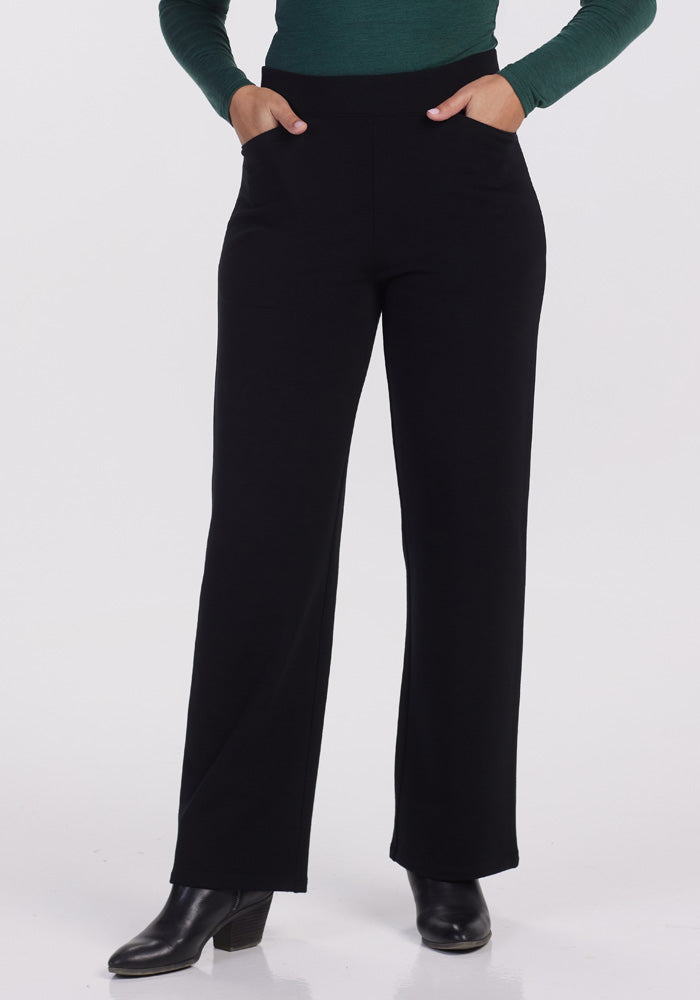 A person is showcasing the Woolx Ellie Wide Leg Pants in black, paired with a dark green long-sleeve top and accessorized with hands tucked comfortably into the pants' pockets. Their look is completed with black ankle boots, all set against a plain white background that highlights the sleek ensemble.