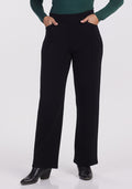 A person is showcasing the Woolx Ellie Wide Leg Pants in black, paired with a dark green long-sleeve top and accessorized with hands tucked comfortably into the pants' pockets. Their look is completed with black ankle boots, all set against a plain white background that highlights the sleek ensemble.