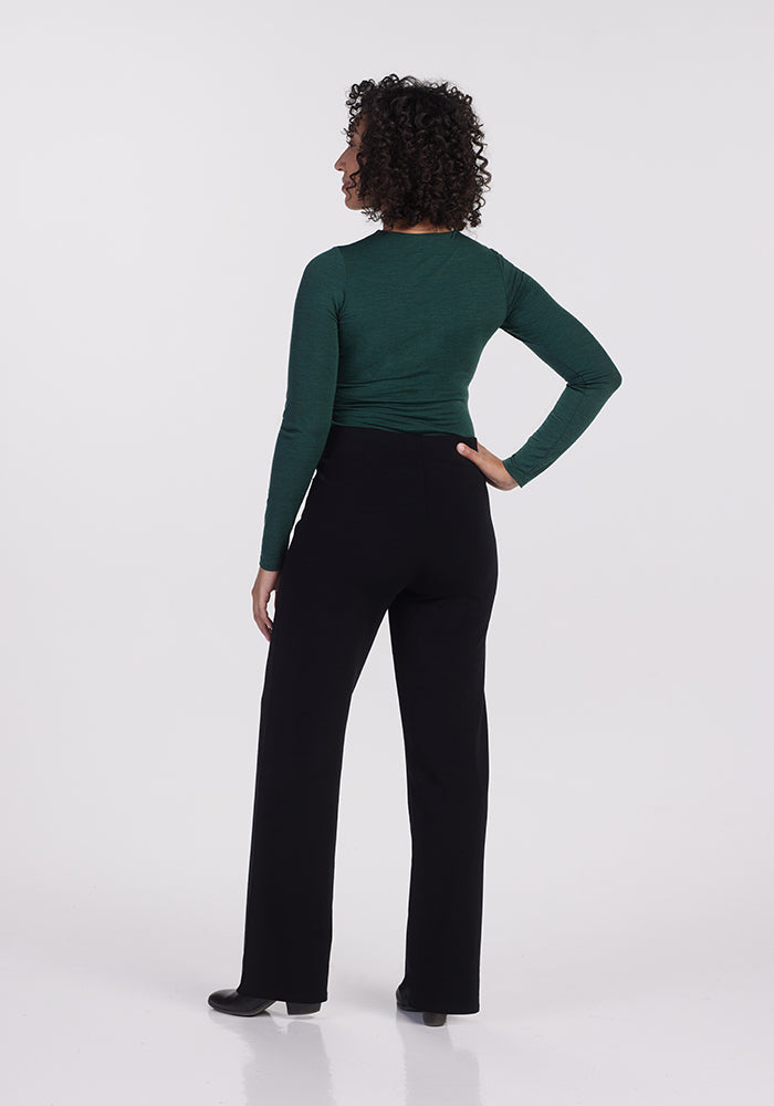A person with curly hair stands facing away, wearing a long-sleeve green top and Woolx's Ellie Wide Leg Pants in black. The plain white background accentuates the simplicity and elegance of the outfit, with the Merino wool ensuring these pants provide exceptional warmth for any occasion.