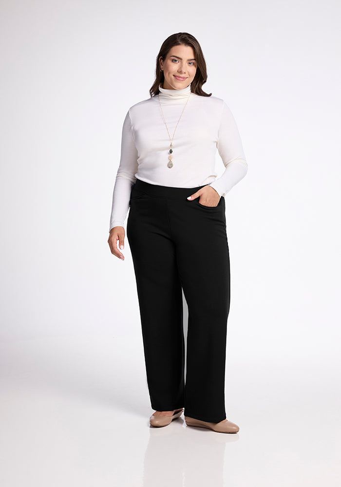 A person wearing a white turtleneck and black Ellie Wide Leg Pants from Woolx, along with beige shoes, stands smiling with one hand in a pocket against a plain white background. They have long brown hair and are accessorized with a long pendant necklace.