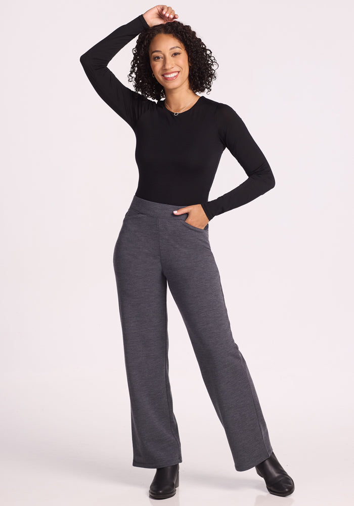 A woman with curly hair smiles, posing with one hand on her head and the other in her pocket. She wears a black long-sleeve top, Woolx Ellie Wide Leg Pants Petite in Cinder Melange, and black boots against a plain light background.