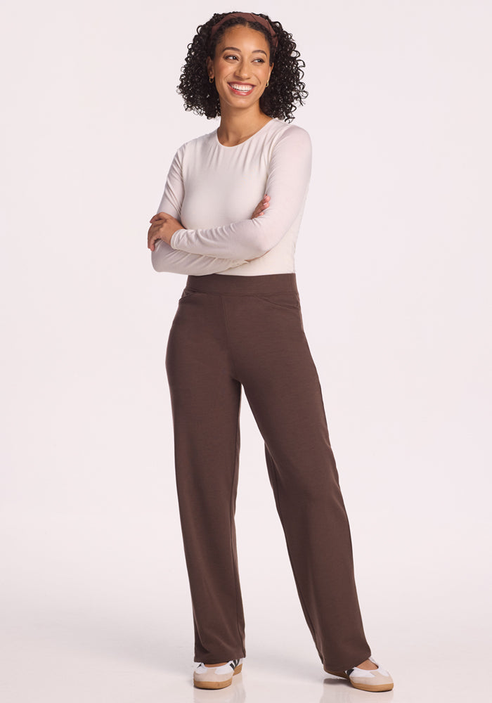A woman with curly hair smiles with arms crossed, wearing a long-sleeve white top, Woolx Ellie Wide Leg Pants Petite in Bracken (Final Sale), and white sneakers against a plain white background.