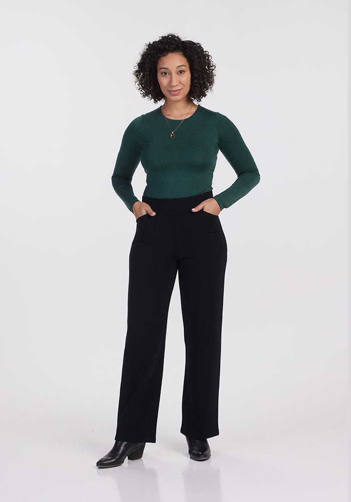 A person with curly hair is standing against a plain background. They wear a dark green long-sleeve top and stylish Ellie Wide Leg Pants - Petite by Woolx, complemented by black shoes. With hands in their pockets and a smile, they exude confidence.