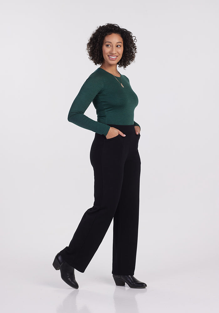 A woman with curly hair is smiling and posing sideways in a green long-sleeve top and the Ellie Wide Leg Pants - Petite from Woolx, made of black Merino wool. Her hands are in her pockets, highlighting the chic wide-leg fit, and she's wearing black ankle boots against a plain white background.
