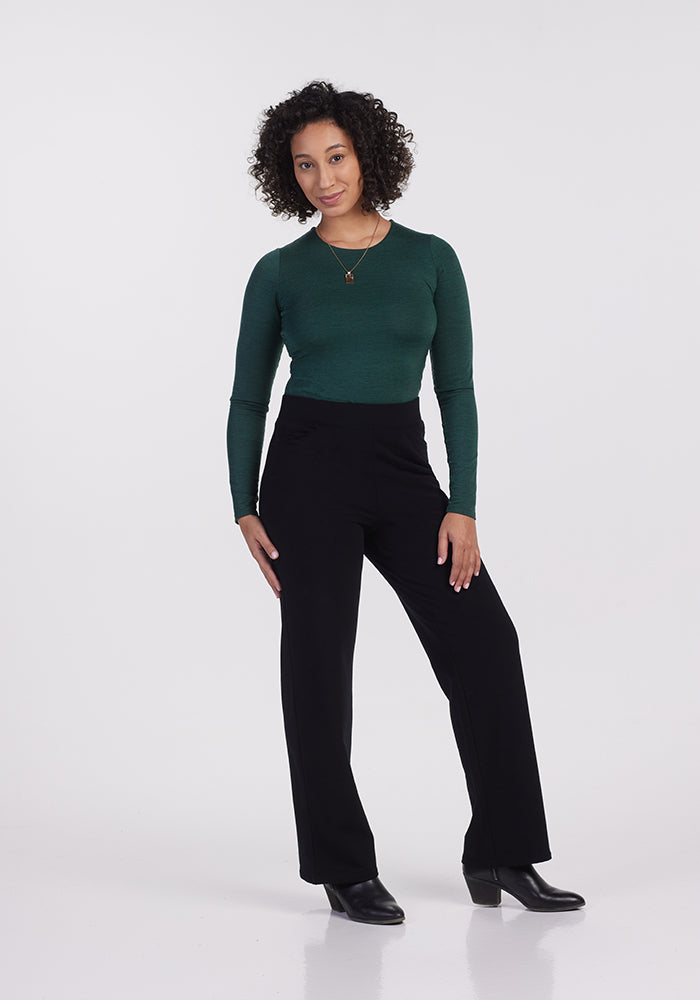 A person with curly hair stands confidently against a plain white background, embodying effortless style in a dark green long-sleeve shirt and the Ellie Wide Leg Pants - Petite by Woolx in black merino wool, perfectly complemented by black boots.