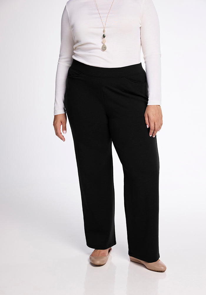 A person wearing a white long-sleeve top, the Ellie Wide Leg Pants - Tall by Woolx in black, and beige shoes stands against a plain backdrop. They are accessorized with a long pendant necklace.