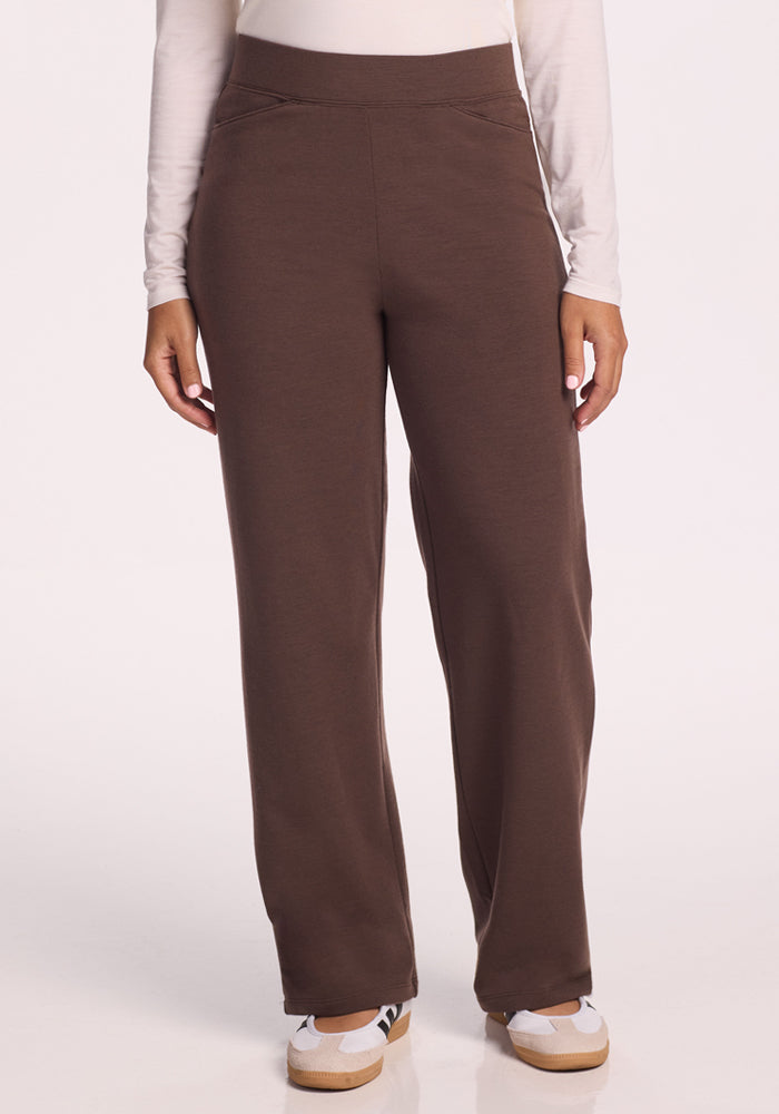 A person stands with arms relaxed, wearing a long-sleeve white shirt and Woolx Ellie Wide Leg Pants in Bracken. White sneakers complete the look against a plain, light background.