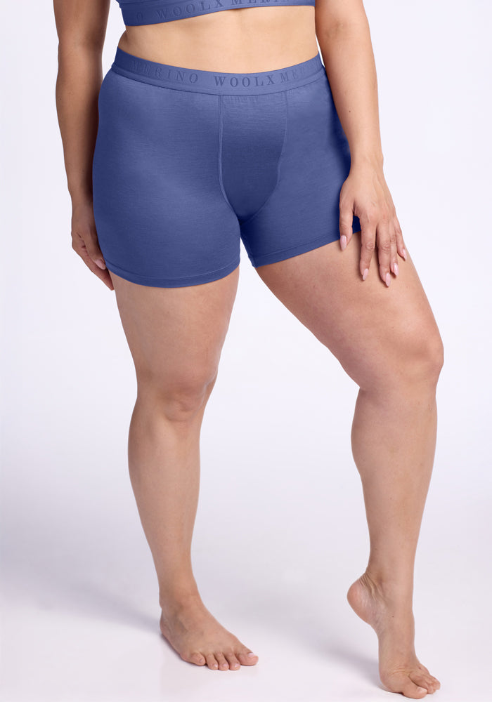 A person wears the Woolx Elliott Boyshort in Cloudy Bay with a matching blue sports bra, standing barefoot against a light background and resting one hand on their thigh. | Juliana is 5'10", wearing a size XL