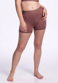 A person stands barefoot against a plain white background, wearing the Woolx Elliott Boyshort in Nutmeg and a matching brown sports bra, with one hand on their hip and the other by their side. | Ariana is 5'11", wearing a size S