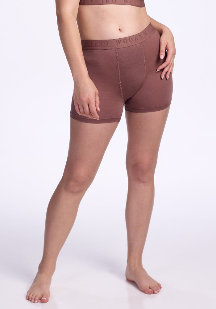 A person stands barefoot against a plain white background, wearing the Woolx Elliott Boyshort in Nutmeg and a matching brown sports bra, with one hand on their hip and the other by their side. | Ariana is 5'11", wearing a size S