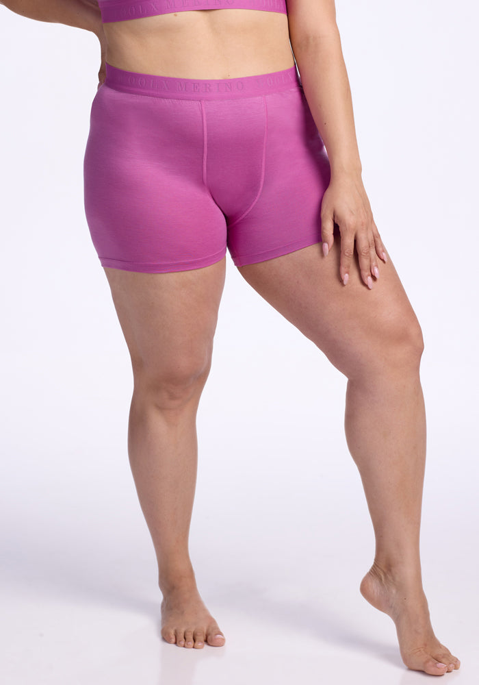 A person stands barefoot on one leg against a plain white background, wearing Woolx’s Elliott Boyshort - Wild Blossom and a matching pink sports bra. Their left hand rests gently on their thigh. | Juliana is 5'10", wearing a size XL