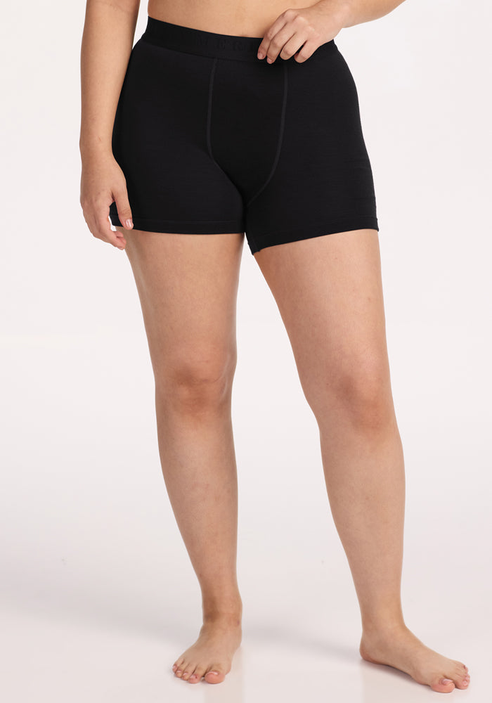 A person stands barefoot against a plain background, wearing the Woolx Elliott Boyshort in black and a matching top, with one hand on their hip. Their face is not visible. | Sarah is 5'8", wearing a size S