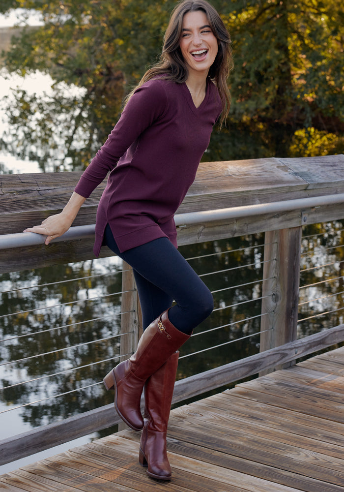 A woman stands on a wooden bridge, smiling and laughing. She wears a maroon sweater, knee-high brown boots, and Woolx Ellis Seamless Petite Tights in Black. Trees and water create a cozy backdrop for this stylish base layer look.