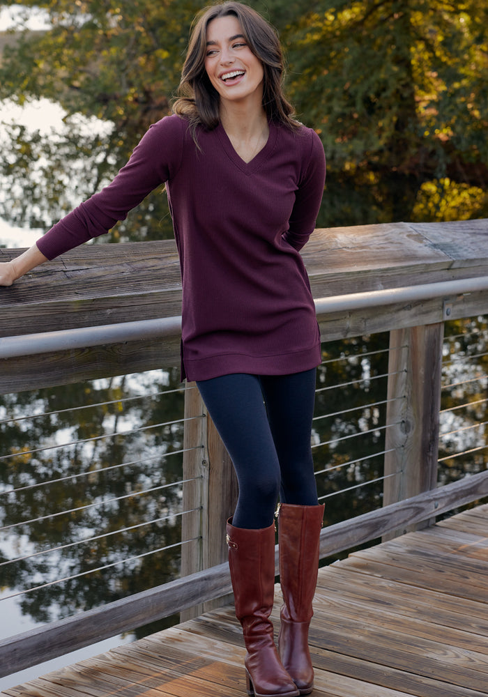A woman with long dark hair, wearing a burgundy long-sleeve top, Woolx Ellis Seamless Tights in black, and tall brown boots, stands smiling on a wooden bridge with trees and water in the background.