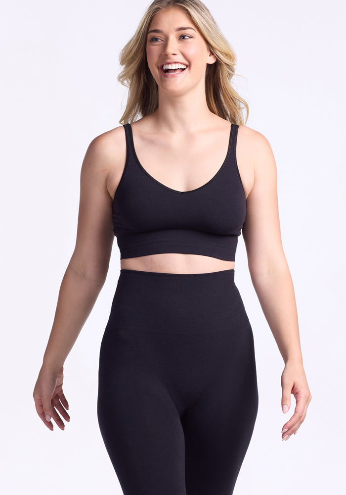 A smiling woman with long blonde hair wears Woolx Ellis Seamless Tights in black and a matching sports bra, standing against a plain light background.