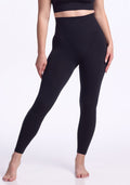 A person stands barefoot against a plain white background, wearing Woolx Ellis Seamless Petite Tights in black and a matching crop top, with their arms relaxed at their sides.