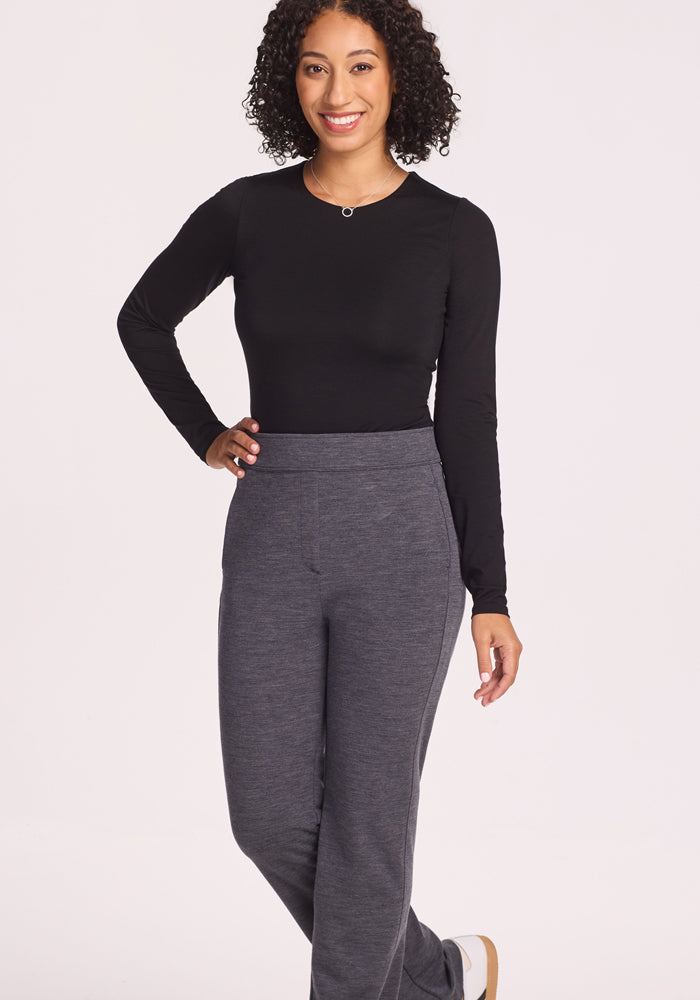 A woman with curly hair smiles, wearing a long-sleeve black top and the Woolx Emerson Straight Leg Pants in Cinder Melange, standing against a plain light background with one hand on her hip.