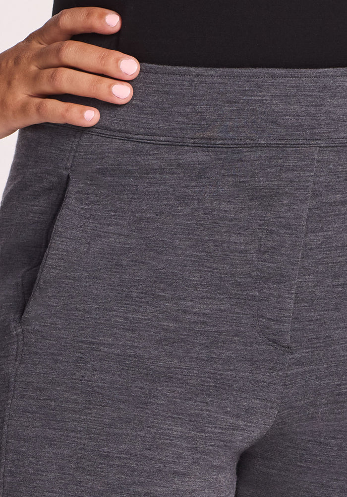 A close-up shows a person in Woolx Emerson Straight Leg Pants Petite - Cinder Melange, their left hand with manicured nails on their hip. The dark gray Merino wool pants, paired with a black top, feature moisture-wicking fabric for style and comfort.