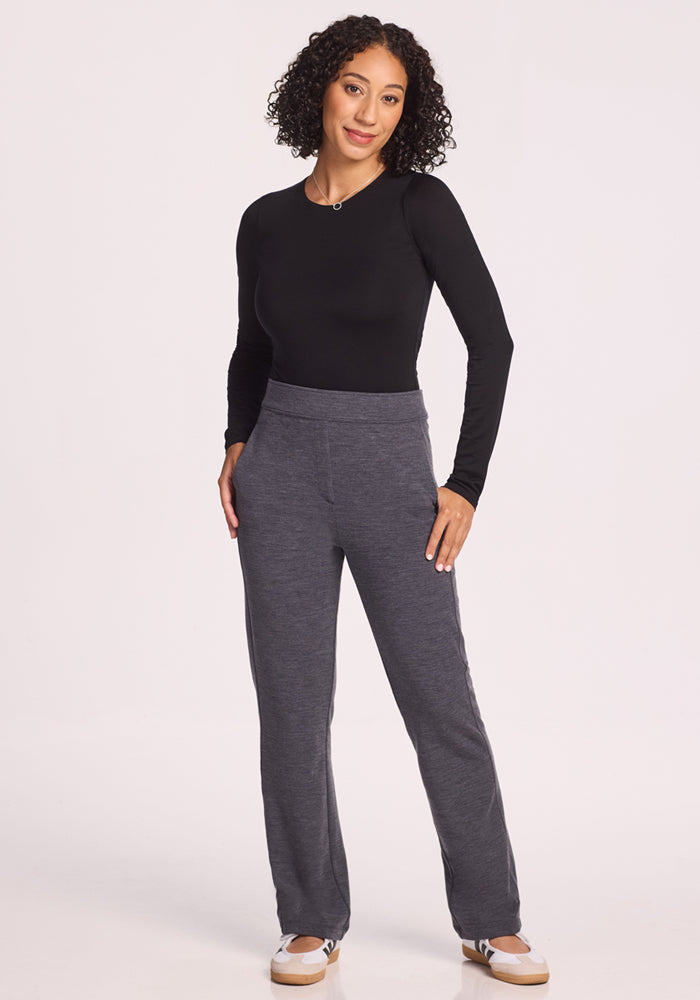 A woman with curly hair wears a black long-sleeve top, Woolx Emerson Straight Leg Pants Petite in Cinder Melange, and beige sneakers. She stands casually against a plain white background, smiling with one hand in her pocket.