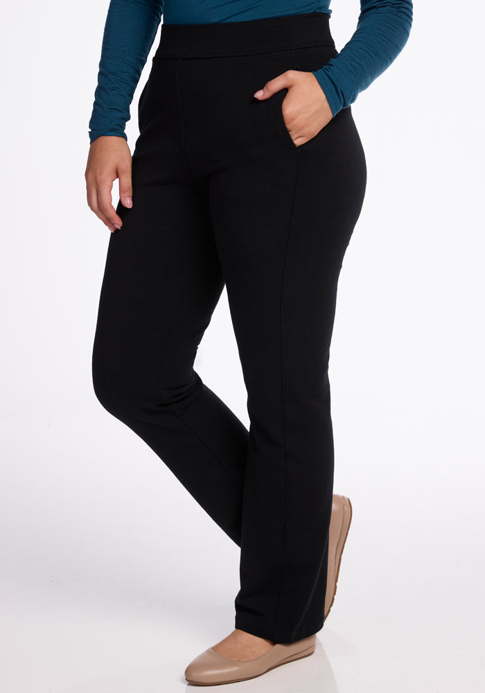 A person is wearing Woolx's Emerson Straight Leg Pants in black, paired with a long-sleeve teal top, their right hand casually resting in a pocket. With beige shoes completing the look, they exude comfort against the plain white background.
